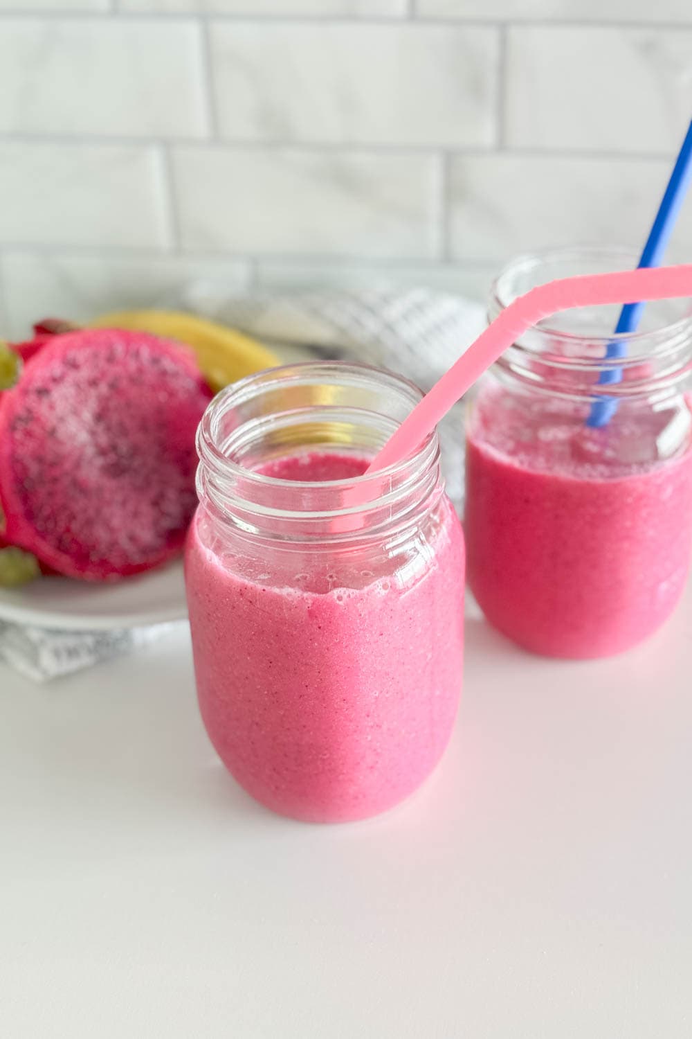 Two mason jars filled with a vibrant pink smoothie that is a vegan tropical smoothie, each with its own colored silicone straw and half a dragon fruit in the background