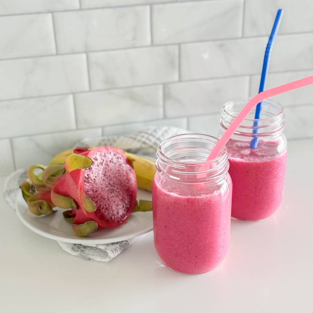 Two mason jars filled with a vibrant pink smoothie that is a vegan tropical smoothie, each with its own colored silicone straw and half a dragon fruit in the background