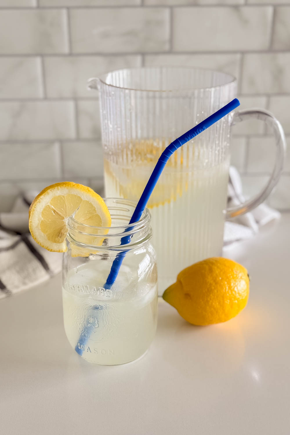 A glass of fresh homemade lemonade next to a full pitcher, with lemon slices and a full lemon