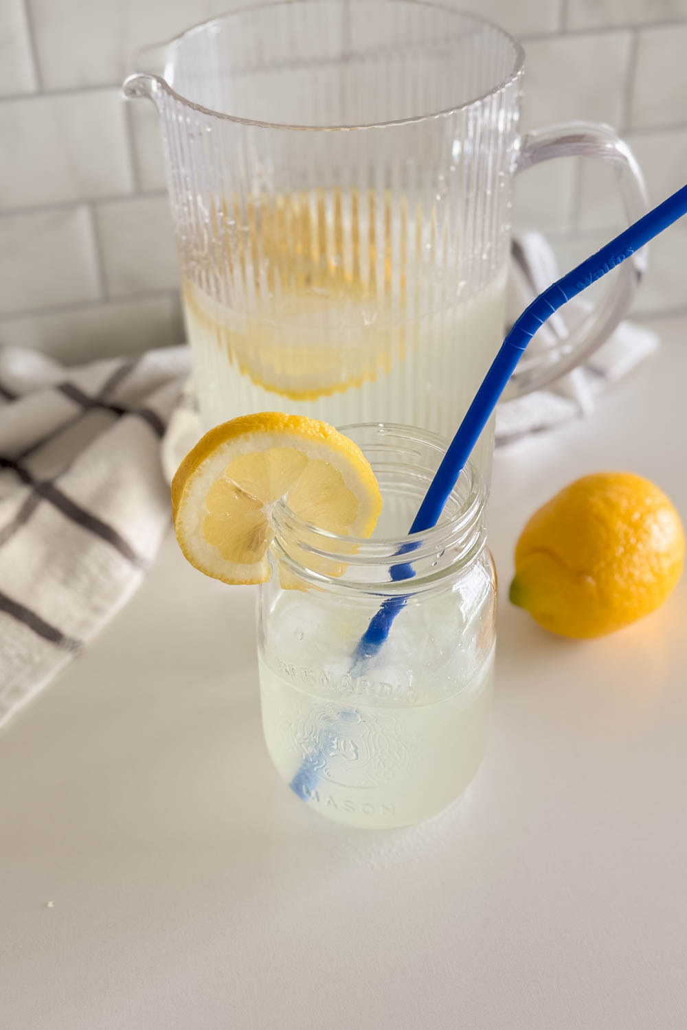 A glass of fresh homemade lemonade next to a full pitcher, with lemon slices and a full lemon